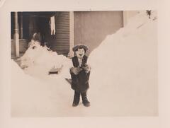 A laughing child standing in the the snow in front of a house. He is wearing what appears to be an oversized vest, hat, and gloves. A pile of snow dominates the right side of the image and there are sleds on the ground behind him.