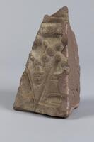 Triangular stone architectual fragment. One side is decorated with carvings creating round and V-shaped elements.