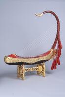 A Burmese harp painted red and black with red tassels and gold inlay. Rests on a gold and red stand.
