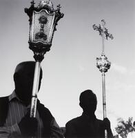 This photograph depicts a view of two men, their faces in shadow, holding Christian processional objects, one bearing a cross.    