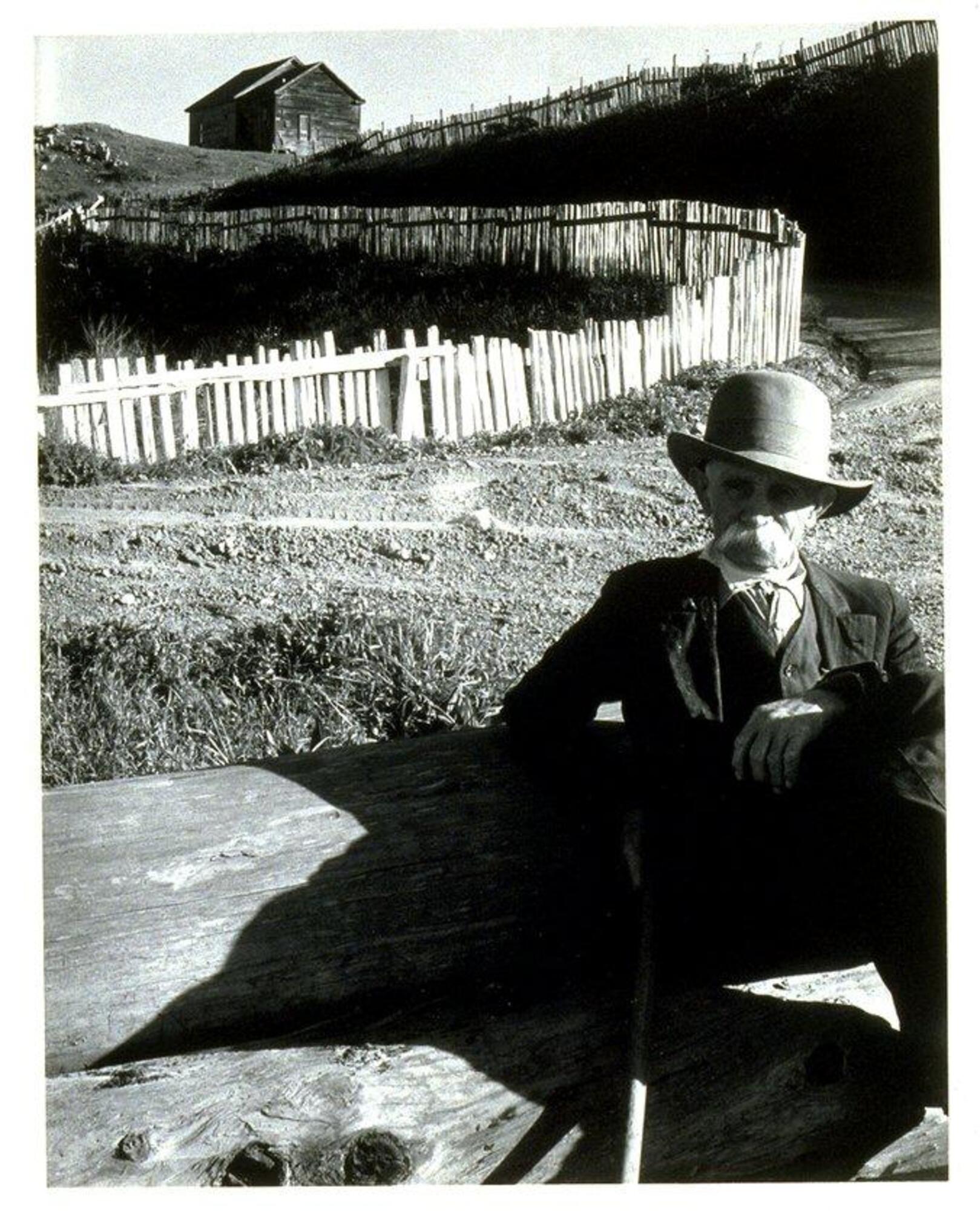 A photograph of a man resting on two large fallen trees. He has a large moustache and wears a casual suit and a hat. 