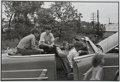 A photograph of four young men sitting in a car. A young child runs past the car in the foreground, blurred from motion.