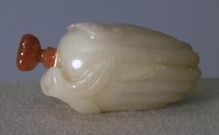 A white nephrite jade snuff bottle, lying on its side in the shape of a citron&nbsp;fruit. At the top of the snuff bottle is an orange glass cap in the shape of the handle. There are also high relief carvings of leaves at the top. Along the body are lines that form curves at the bottom.