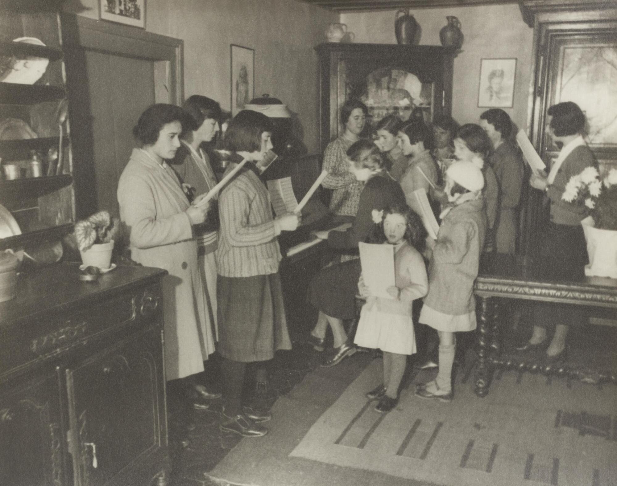 Interior view of a group of women holding sheet music and a single woman sitting at a piano. A hutch is on the left side of the photograph.