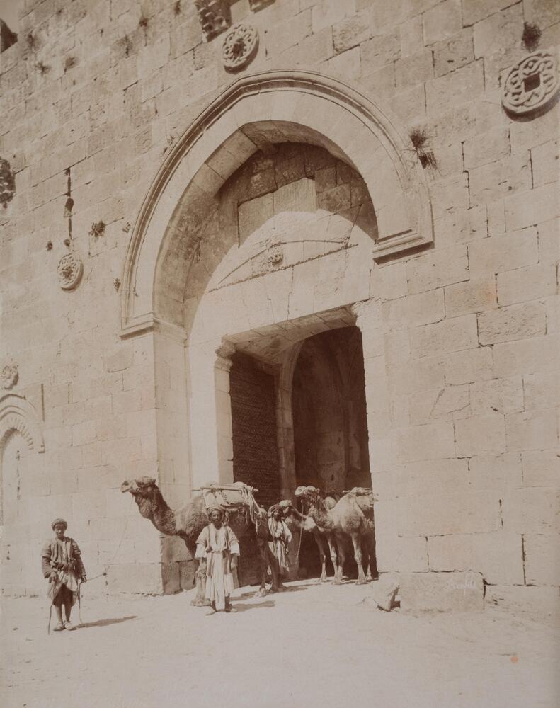 Three male figures and three camels stand in or near the threshold of a stone portal. The three men face the viewer while the camels are in three-quarter turns facing the left-hand edge of the photograph.