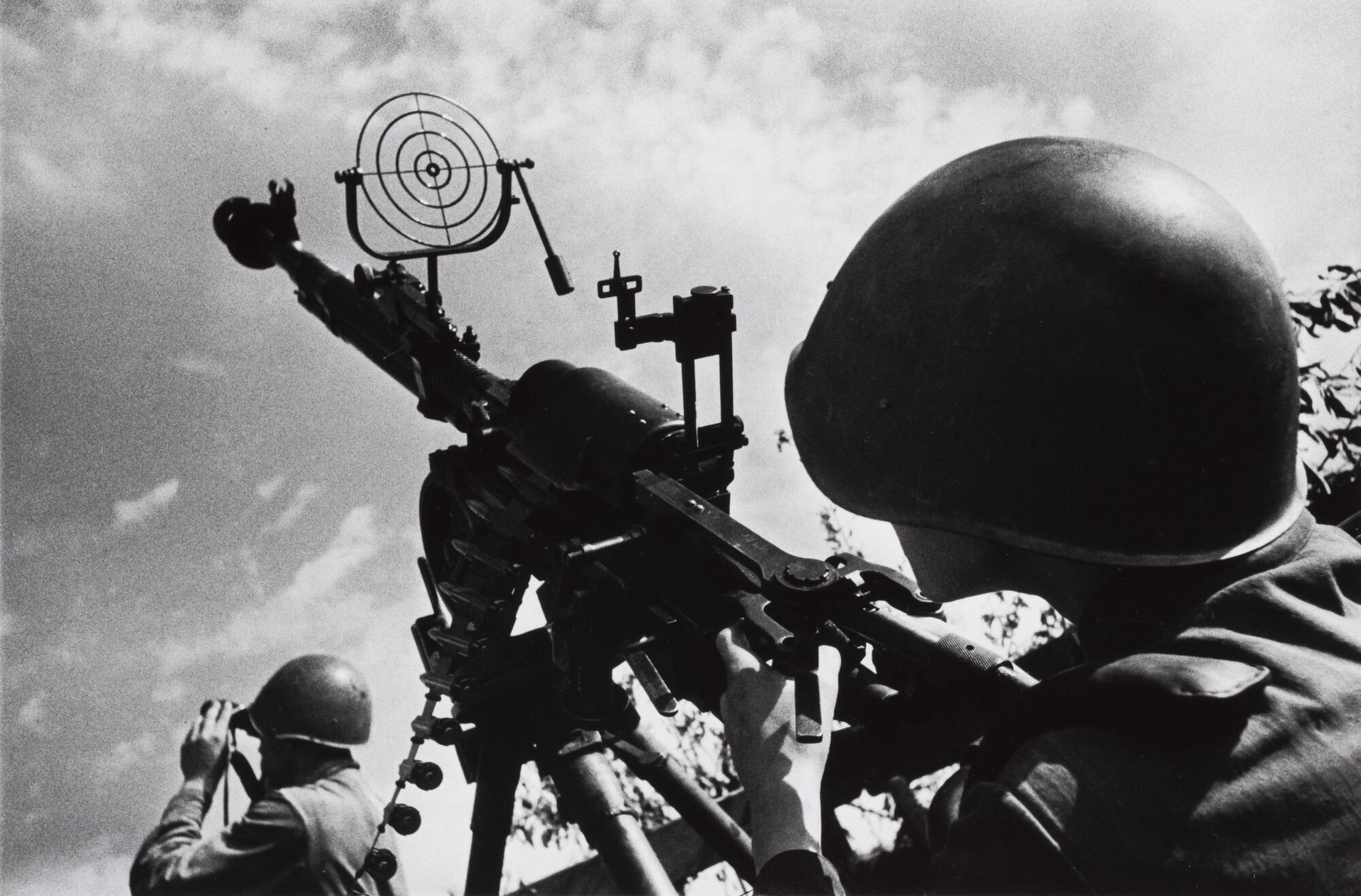 View down the barrel of an anti-aircraft gun seen from behind the shoulder of a young soldier; another soldier with binoculars is visible in the lower left.