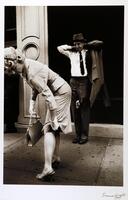 This is a black and white photograph that shows a woman and a man standing on a sidewalk in front of a doorway. The woman, closest to the viewer and shown in profile, is bending over and reaching behind to adjust her skirt. The man is standing with arms raised to adjust his collar before putting on his sportcoat.