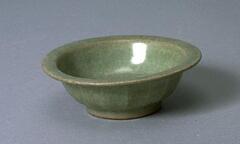 A small stoneware bowl on a foot ring with an everted flat rim. The exterior is carved to represent lotus petals, the interior molded with two fish. The bowl is covered in a green craqueleur celadon glaze.