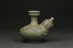 A green vessel, or kendi, with a flat oval body with a wide spout on the side that sharply thins at the tip. The neck at the top of the kendi&#39;s body is long and suddenly widens at the rim. The outside is decorated with geometric designs.<br />
<style type="text/css"><!--td {border: 1px solid #ccc;}br {mso-data-placement:same-cell;}-->
</style>
