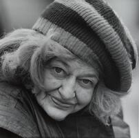 A close-up portrait of an older woman with light hair and a knitted striped cap. She is looking ahead and smiling.