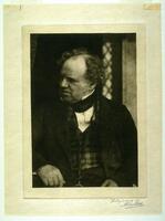 This photograph is a portrait of an older, seated man in a three piece suit. He is looking to the side in an interior space.