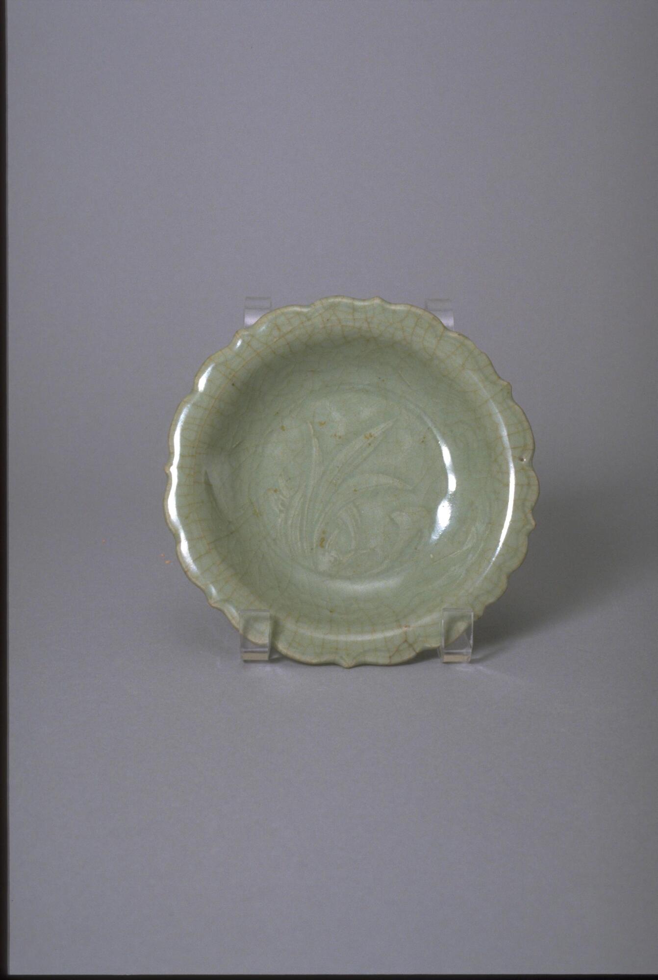 This stoneware shallow bowl has a flat bottom on a foot ring with everted molded eight-panel foliate rim. The interior is incised with plant design and covered in a green celadon glaze with craquelure finish. 