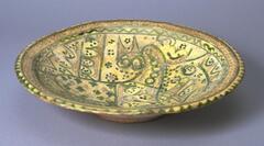 This large Iranian plate sits on a small base. The decoration consists of a bird and crude space fillers. The units are outlined in green and incised, in addition to green glazed parts that are not incised. The design is cut through the slip and appears light brown while the exterior is left unglazed and plain red-brown.The plate is mended and appears to have commercial mosaic in parts. The colors are green, light green, yellow, and red-brown. Painted green and brown spots and stripes are combined with scribbled engraving to form often asymmetrical designs.