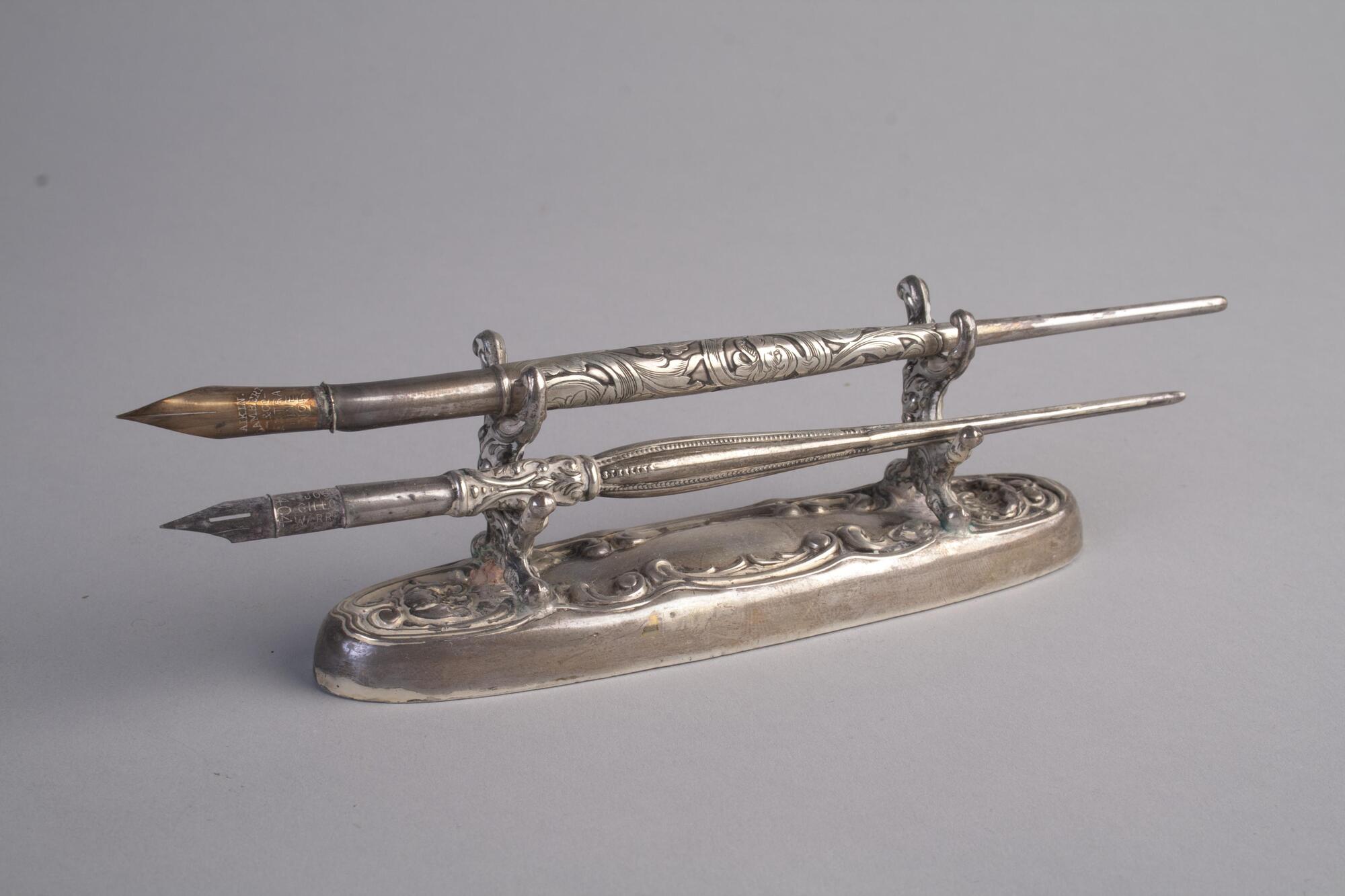A pen holder with two pens. Both the pens and the holder are sterling silver and have scroll patterns.