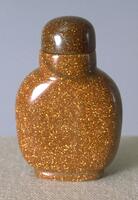A golden glass snuff bottle with copper flecks that resemble glitter. On the top of the snuff bottle is a brown glass snuff bottle with copper flecks.