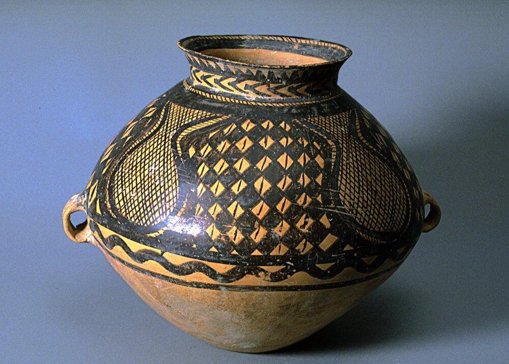 A light reddish-buff earthenware <em>guan (</em>罐) jar with a wide globular upper body and conical lower body on a narrow flat base, a tall wide neck and a flaring rim. Two diametrically opposed lug handles are located at the waist. The upper half of the body is painted with black pigment to depict four gourd-shaped areas containing a network pattern. The negative space between the gourd-shaped areas contains a checkerboard pattern confined between solid band borders, with a lobed line border below, and one final solid black band at the bottom of the design. Around the neck are bands of herring bone, sawtooth, and hatch mark patterns, and additional hatch marks and solid bands toward the interior.  