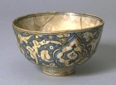 This deep footed bowl comes from the 17th century Safavid period in Iran. The bowl features an ivory ground with a glossy glaze and blue and black underglaze painting. Cobalt blue floral rug designs cover the exterior of the bowl and  a cobalt blue medallion is found on the interior base.
