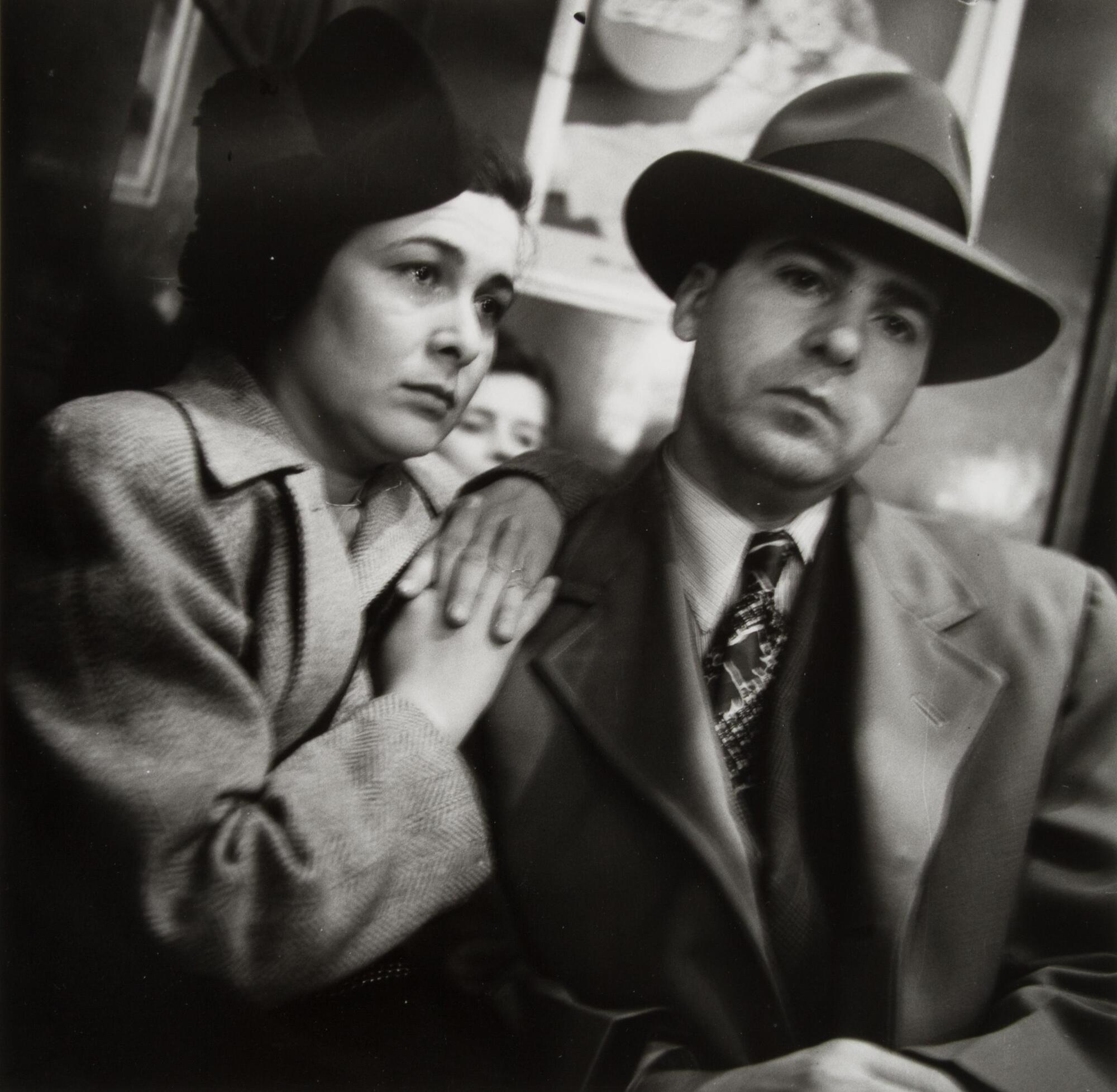 A man and woman seated. Her hands are folded together, resting on his shoulder. He has his hands on his lap, wearing a suit, tie, hat and overcoat. Behind them is a Coke sign that is blurry.