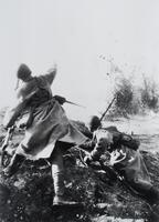 Back view of two soldiers on a dirt mound—one standing with arm outstretched, the other crouching down—with an explosion in the distance. 