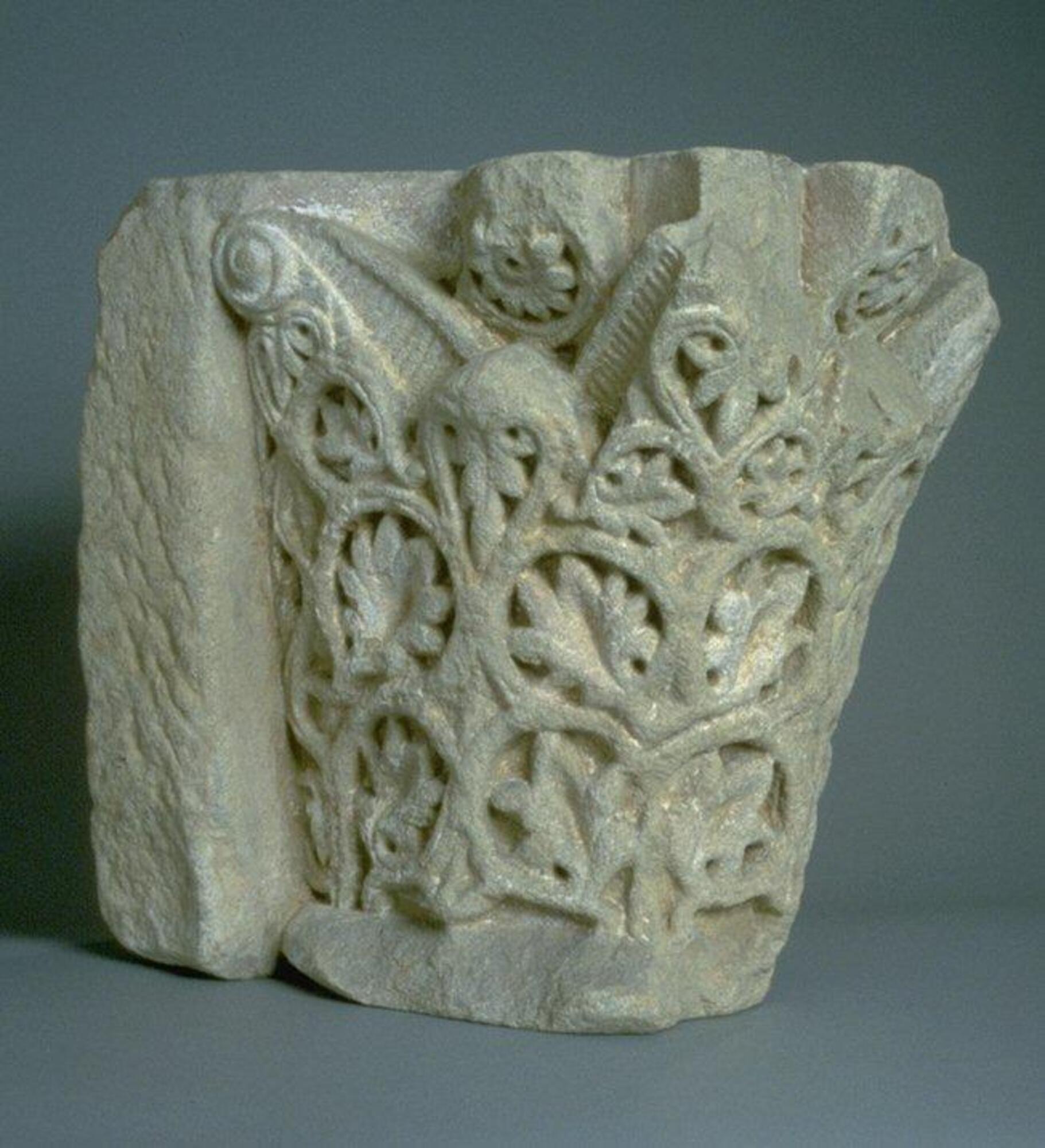Engaged capital carved in grayish, coarse sandstone (arkose). The bell-shaped drum is decorated with a pattern of vine rinceau that encircles palmette leaves in a roughly symmetrical arrangement on each face of the capital. These ornamental plant forms are deeply undercut to highlight the pattern in sharp relief. A pair of volutes decorated with vertical striations springs from the vine rinceau in the upper portion of each face of the capital. A rosette enclosed in a circle appears at the top edge of the center of each face of the capital, above the point where the branches of the volutes diverge.