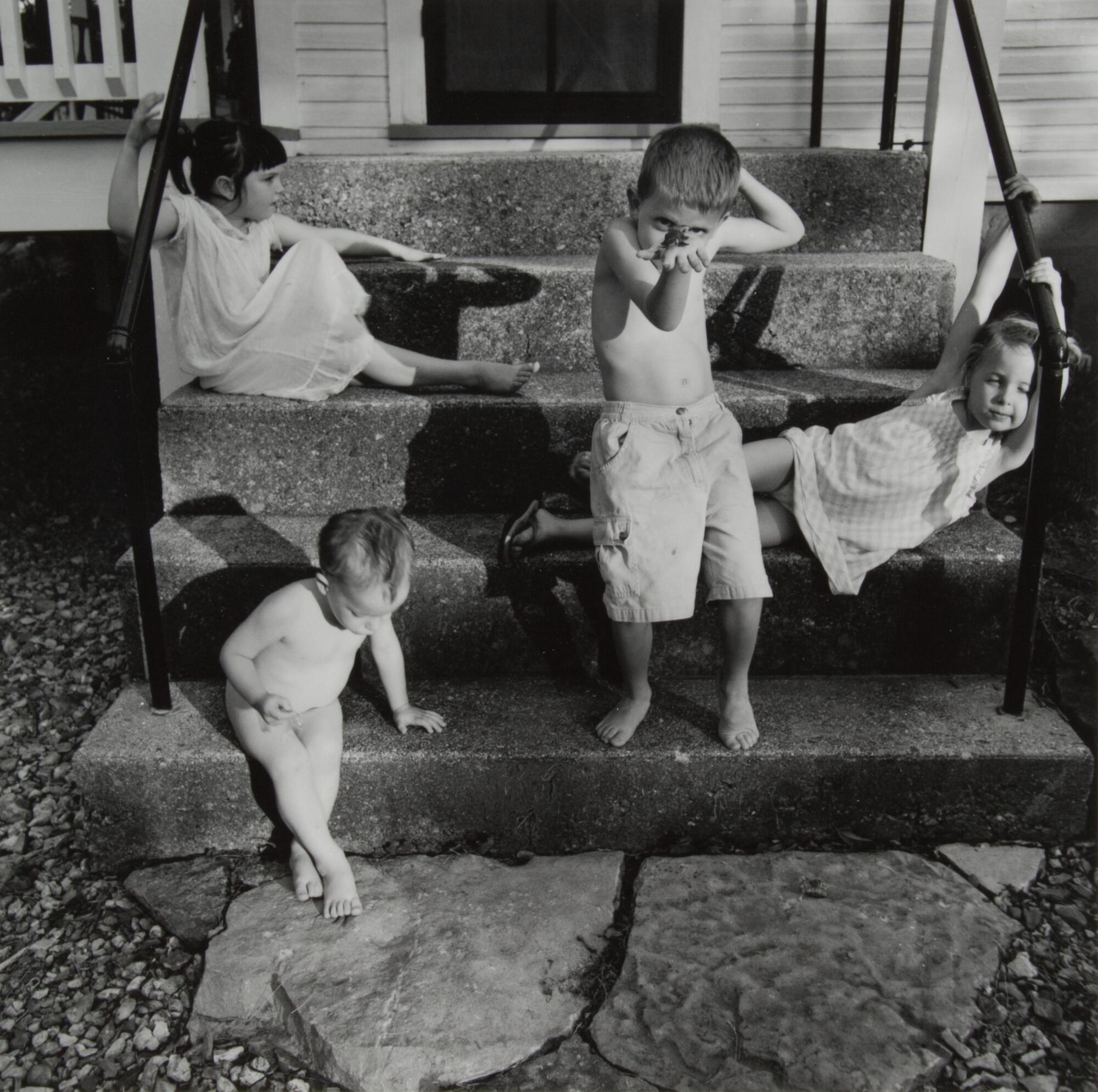 A photograph of four children in front of a house entrance. Two girls hold onto the railing and recline on either side. An infant sits on the bottom step, while another young boy stands on the first step, holding a frog in front of his face.