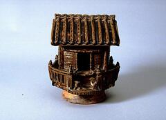 Model of a wooden house with a large roof and a fence surrounding it. On the front deck is a dog. The model is on a large and thick footring.