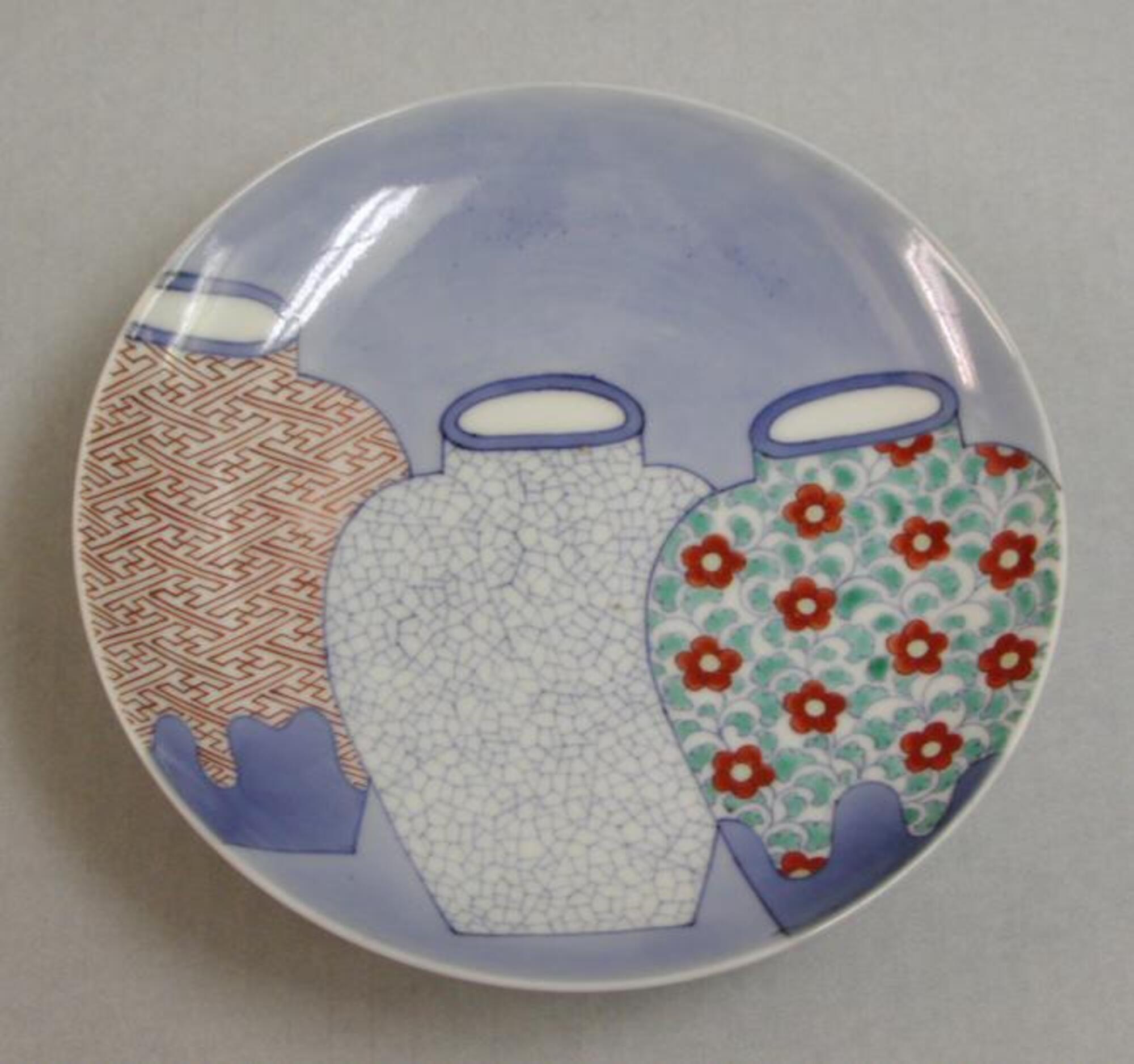 A circular, smaller white porcelain plate. On the upper surface, three vases, similar in shape, are shown overlapping diagonally in blue background. The two outer vases run off the plate’s rim. They are outlined in underglaze blue and are against a painted light-blue background. The left vase is covered with a white glaze (which left unpainted) under a red key-fret design. The center vase is totally covered with a white crackle pattern done in blue underglaze line. The right vase has a design of evenly-spaced red flowers. Precise flat bases are combined with a rather awkward suggestion of a three-dimensional view of the lips. The back has a triple representation of peony buds surrounded by fine branching stems and leaves. On the shallow foot, bold lines are drawn in a row like a comb. The design on the back is all drawn with blue underglaze. (Referencce: Becker, Sister Johanna. “A Group of Nabeshima Porcelain.”)