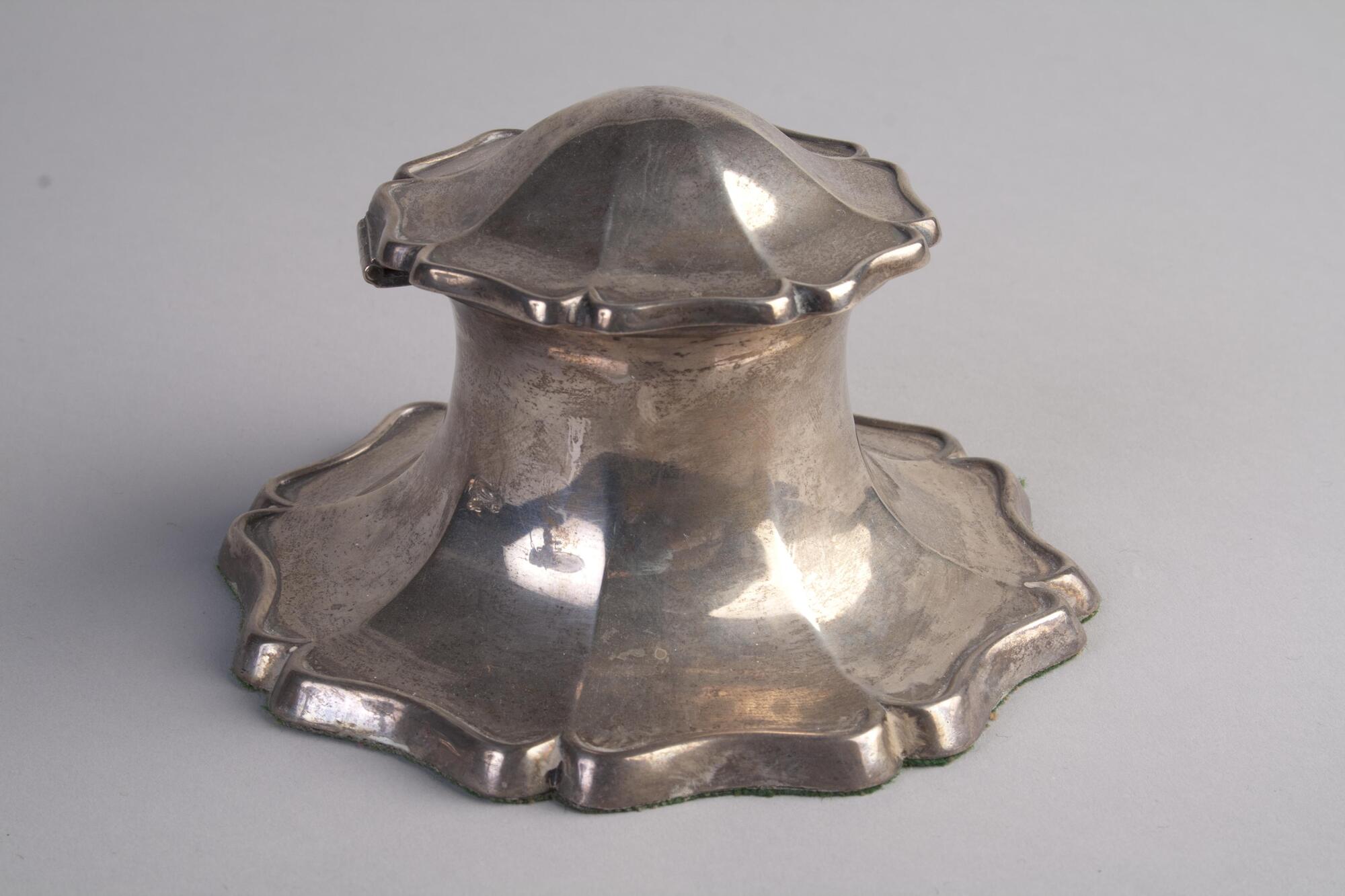 A silver metal inkwell has a shape and body like a foundain. The edges of the bottom and the lid of the inkwell is irregular.