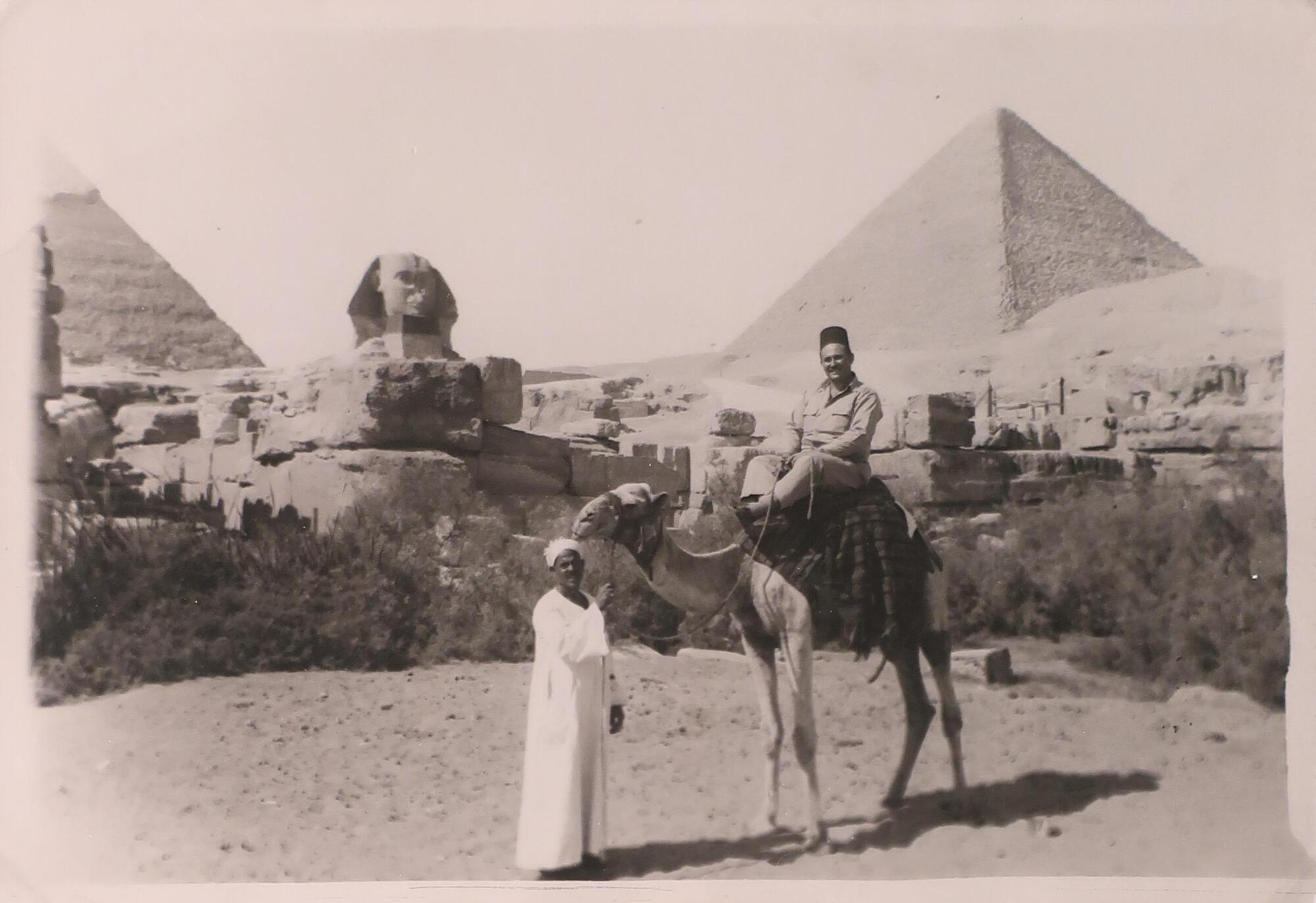 Two men, one standing and another riding a camel, in front of two pyramids and a sphinx statue in the distance.