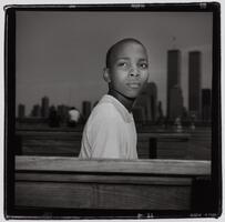 A portrait of a young boy. He turns his head over his shoulder, his lower torso obscured by a wooden structure in the foreground. In the background, a city skyline is visible.