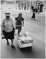 Two women walk along a cobble stoned road. One wears a straw hat and daisy patterned shirt. Ther other wears a dress with roses on a dark background. She pushes a baby wearing light clothes and hat in a light-colored stroller.