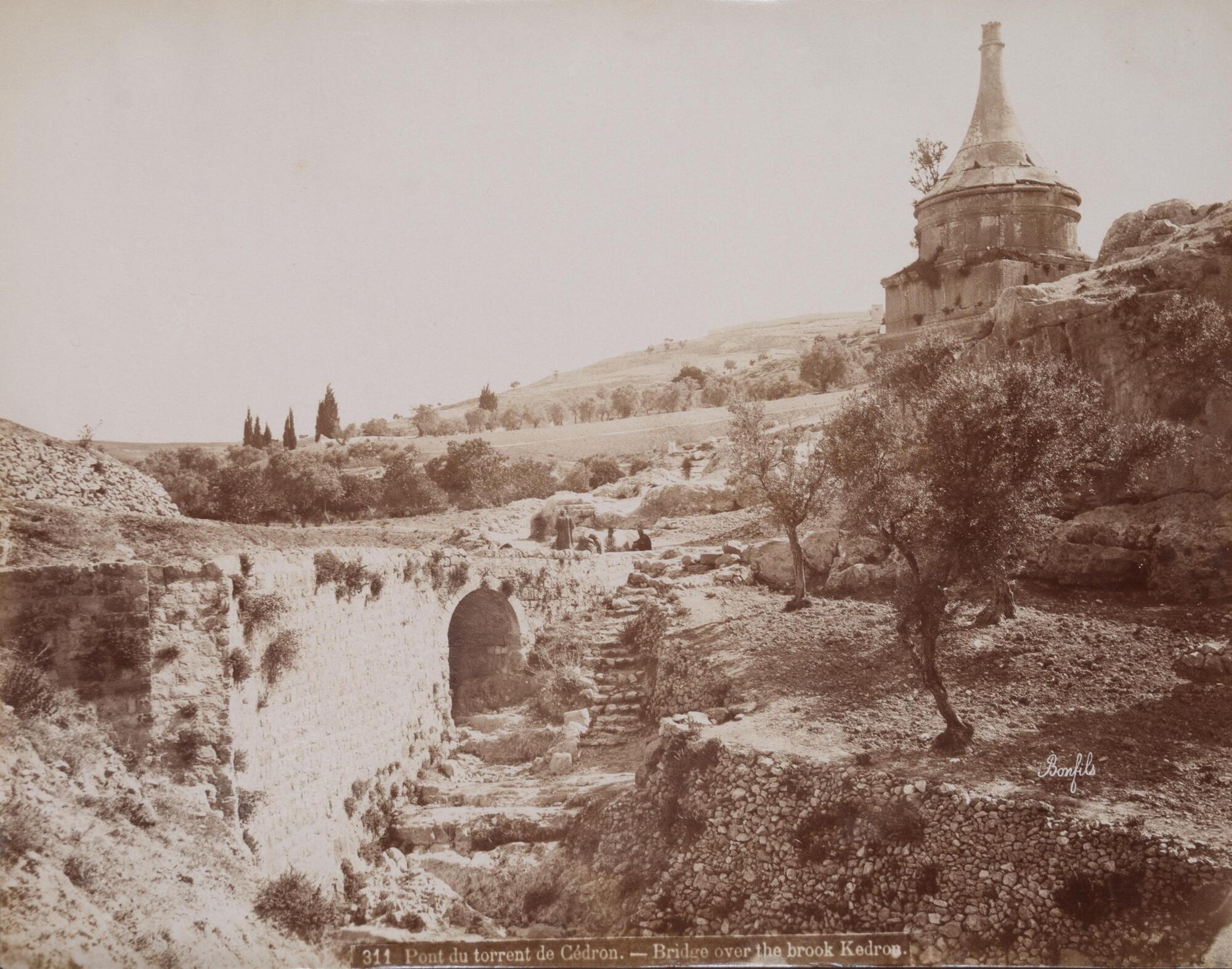 A small stone bridge pierces a countryside landscape otherwise filled with tree-dotted hills. A cylindrical tower dominates the upper-right corner of the photograph. 