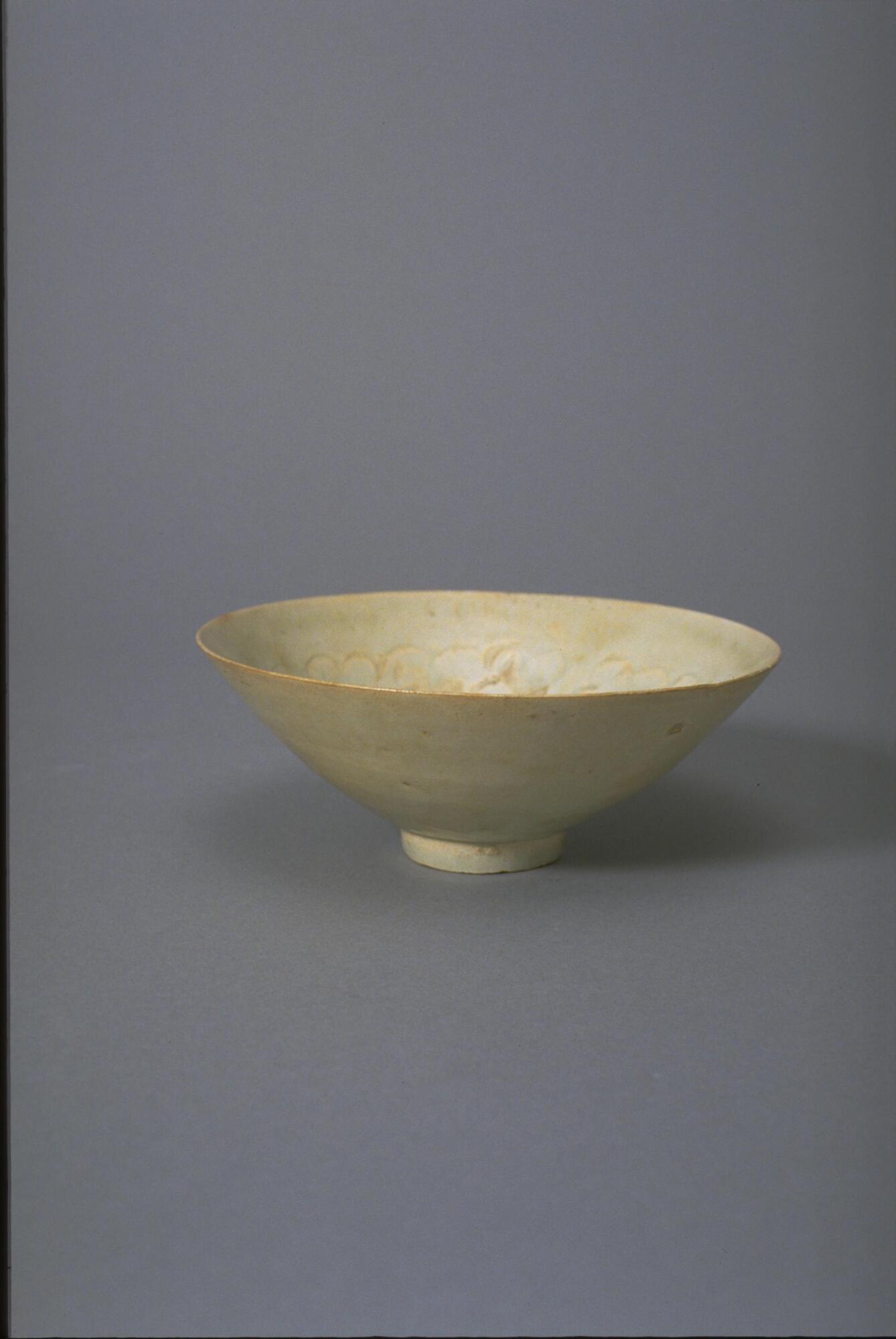 This thin porcelain conical bowl with direct rim on a footring has an interior with incised and combed cloud decoration. It is covered in a white glaze with bluish tinge and has an unglazed rim.