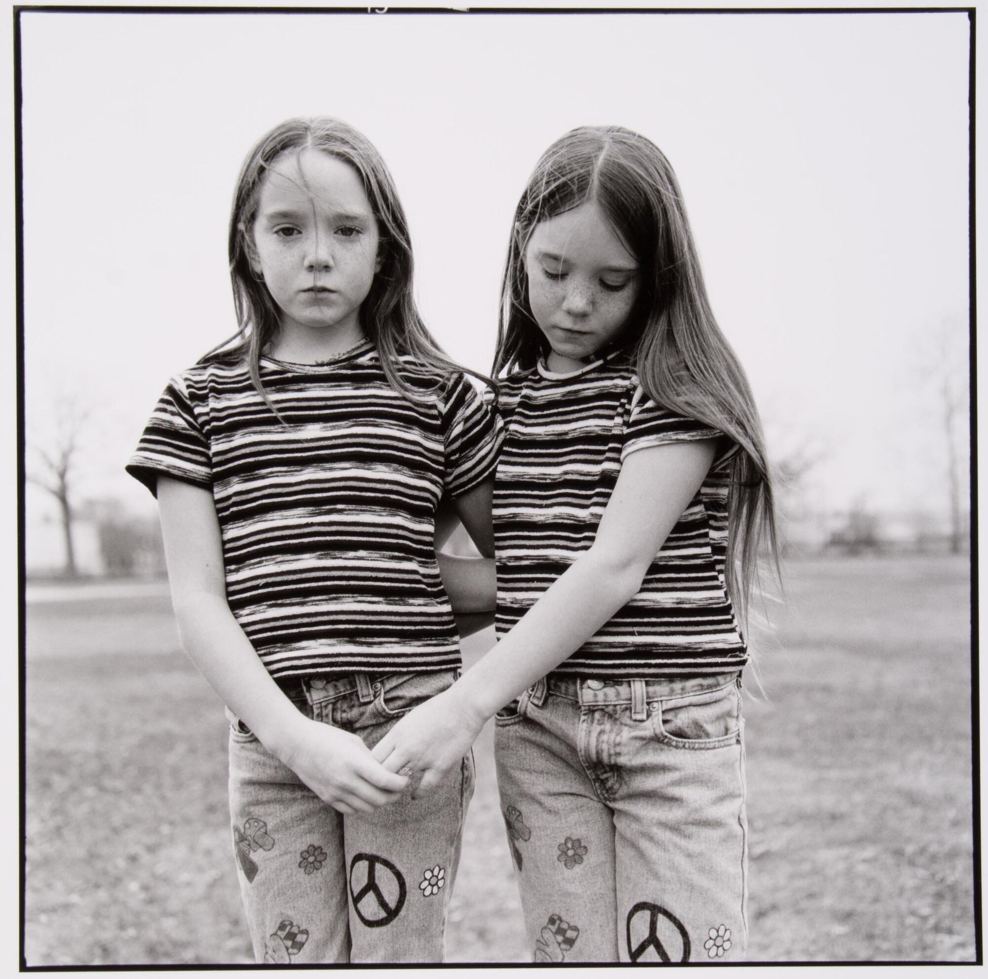 Twin girls wearing matching outfits and standing in a field. They are holding hands and one is looking at the camera, the other is looking down.