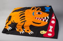 Black background with red trim on either side, but not on the top and bottom. There is a large orange tiger in the center with a red donkey and blue bird on the left side. Colors of blacks, greens, reds, blues, oranges and yellow. There is lilac trim on the top and bottom of the cloth.
