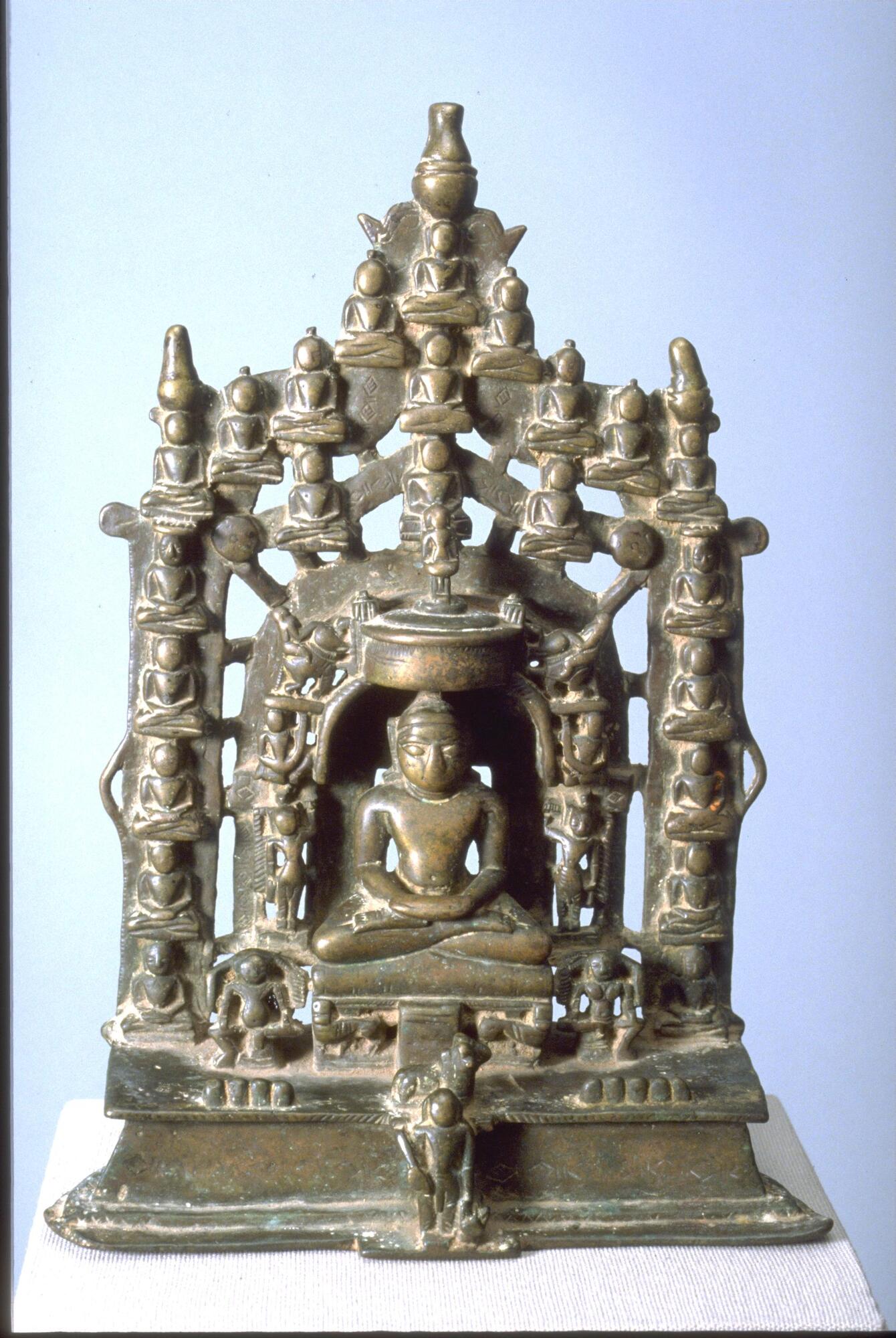 This shrine depicts a large seated Jina surrounded by 23 other jina figures and a variety of attendants.  The Jina figures that adorn the sides and are arranged in tiers above the main figure. The side columns and the whole is surmounted by auspicious pot forms.  The main figure sits in the lotus position on a lion throne flanked by a male and female demigod.  Along the sides he is flanked by standing cauri bearers, garland bearers above them and riders on elephants above that with an umbrella with a standing figure on it above his head.  At the base in the center is a standing figure holding a sick or club with a bull cognizance behind him on the base of the throne.  The nine globs on the base, four to his right and five to his left represent the nine planets and his hands folded in a gesture of meditation<br />