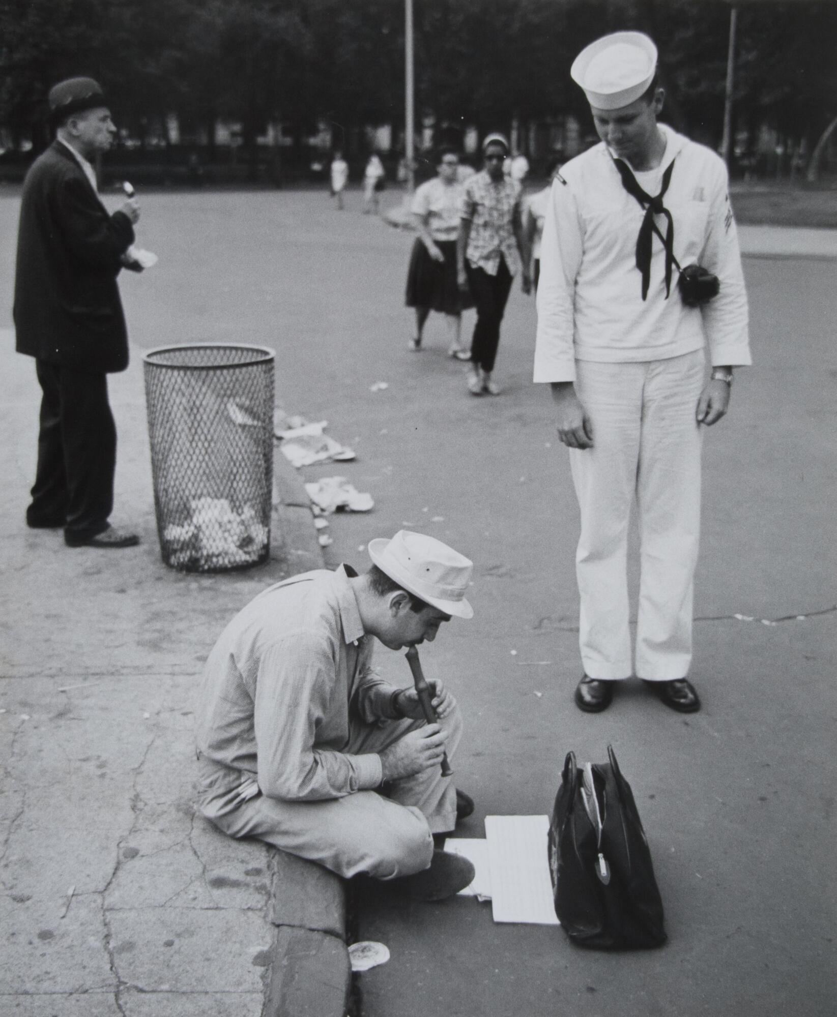 A street scene. There is a man playing the flute and a sailor standing. There is a man in a suit behind him, next to a trash can. There is litter all over the ground.