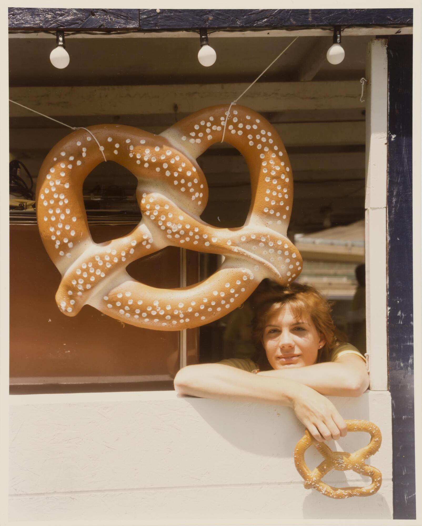 This is an image of a woman at a food stand beneath a suspended sign in the shape of a pretzel.