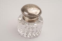 This is a crystal inkwell with a silver-plated cap. The bottle has a cylinder shape with vertical fluting. The round cap has hammered designs on top and a beading pattern around the edge.
