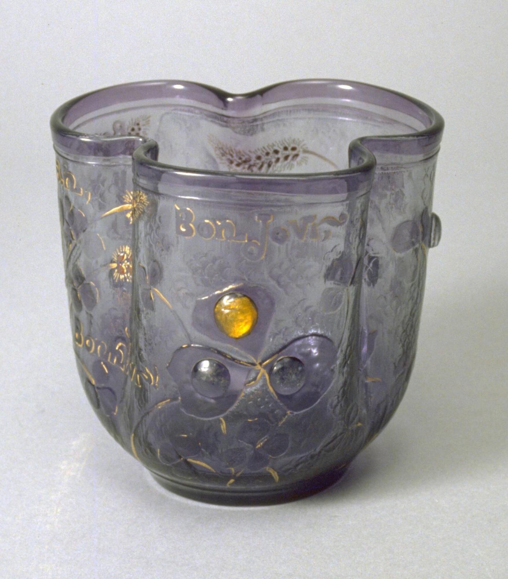 Vessel with trefoil, or three-lobed, shape of transparent blue glass with an intricate plant-motif decoration and gold accents. <br />