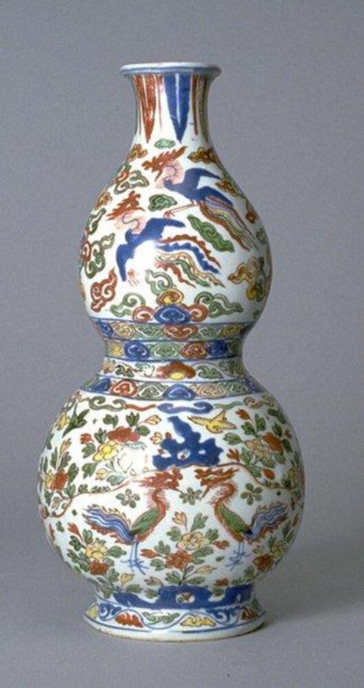 A porcelain bottle vase of double gourd form on a flared footring with tall narrow neck and the reverse side flat with a slot for hanging. The vase is outlined in underglaze blue and has a six-character Wanli mark in a plaque framed by a polychrome overglaze lotus leaf on the top, and a lotus flower on the bottom. The front is decorated with underglaze blue and polychrome overglaze enamels to depict a pair of phoenixes flying among clouds on the upper bulb, and two phoenixes facing each other among an earthly flower garden on the lower bulb. These are confined between<em> lingzhi</em>-shaped clouds and lotus meander borders, with banana leaf lappets around the rim, all covered in a clear glaze. 