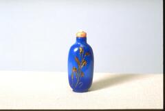 A blue glass oval shaped snuff bottle on a footing. On the front of the snuff bottle is a sparkly gold plant design. At the top of the snuff bottle is a mouthpiece with an orange stopper.