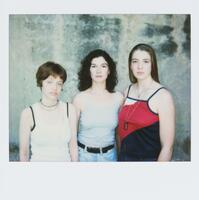 This photograph depicts a chest-level portrait of three young women wearing tank tops and facing the camera. The three women stand against a cement wall.