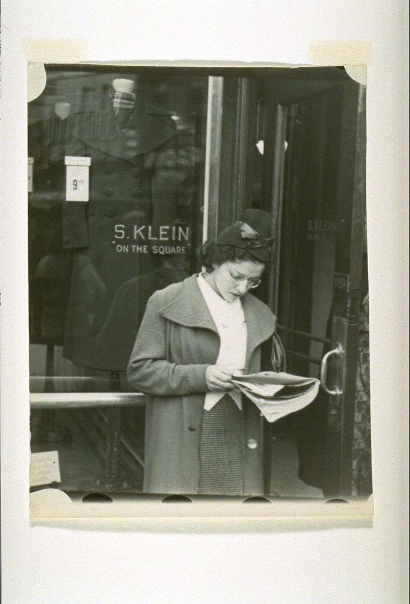 Photograph of a woman reading at a bus stop in front of a department store vitrine.