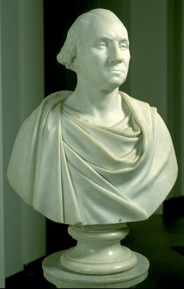 This is a white marble sculpture that depicts the upper torso of a man. There is drapery wrapped loosely around his figure. He is shown staring into the viewer's space and his facial expression is calm and reserved. This sculpture is carved in a realistic manner and rests on a columnar pedestal.