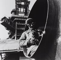 A photograph of a man sitting at the opening of a large pipe. He welds along the pipe's rim.