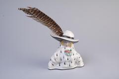 An inkwell made with porcelain has a trianglar shape body and hat-shape lid on top, there is a feather sticking on the hat-shape lid.