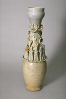A tall porcelain vase with an ovoid lower body, and a tall, narrow ribbed neck, and dish shaped mouth. The jar is on a footring. The neck has several applied figures of molded immortals and sculpted cosmological creatures, including a dragon and clouds.  The jar is covered in a white glaze with a bluish green tinge. It is missing the lid. 