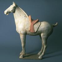 This is an earthenware model of a horse dressed in a saddle blanket and saddle. It has a short, upward pointing tail and trimmed mane. The saddle is painted with reddish mineral pigment. 