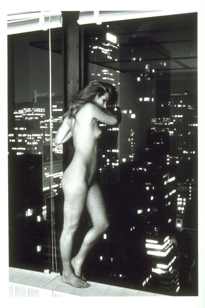 This photograph depicts a nude woman standing in a large window overlooking an evening cityscape.
